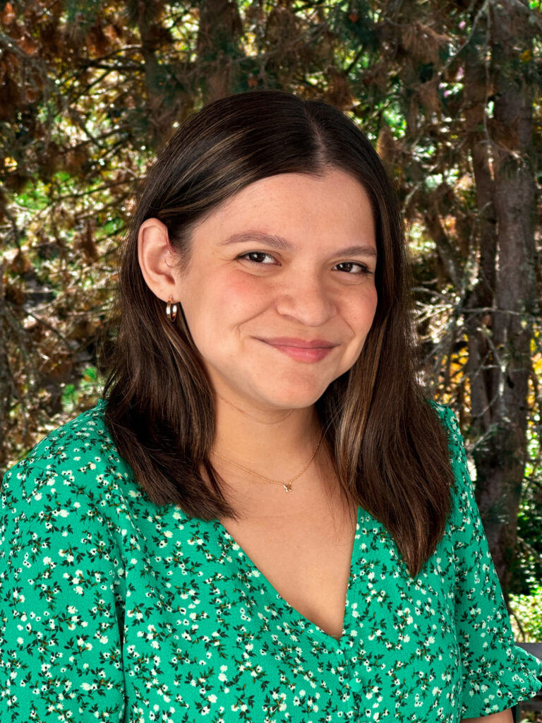 Ana smiling outside, wearing a green blouse with trees in the background.